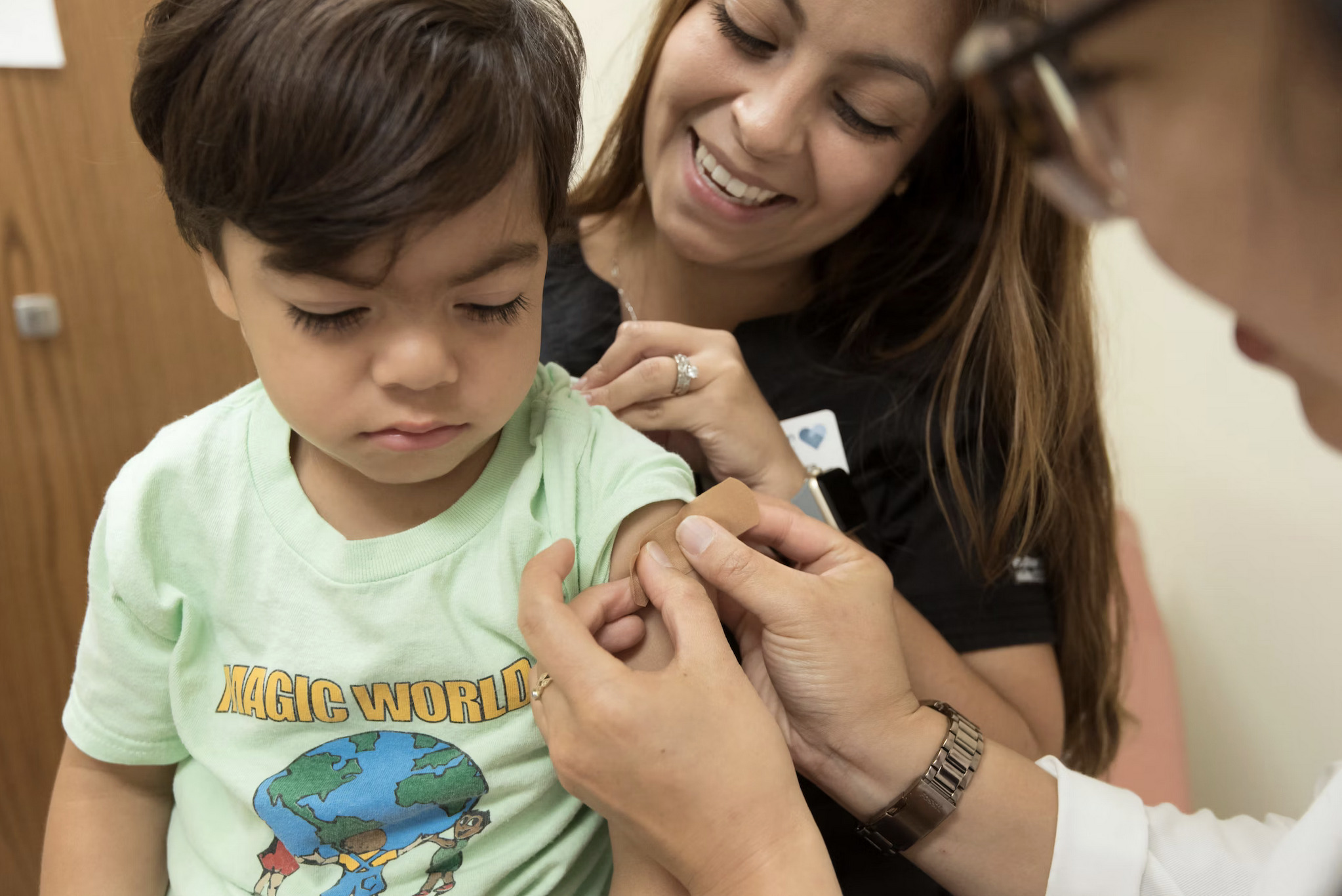 Picture of a young boy who just received a vaccination. He is sitting on his mother's lap and is receiving a band-aid from a healthcare professional.